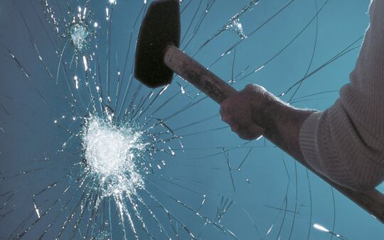 Mann mit Vorschaghammer vor zerstörter Glasscheibe